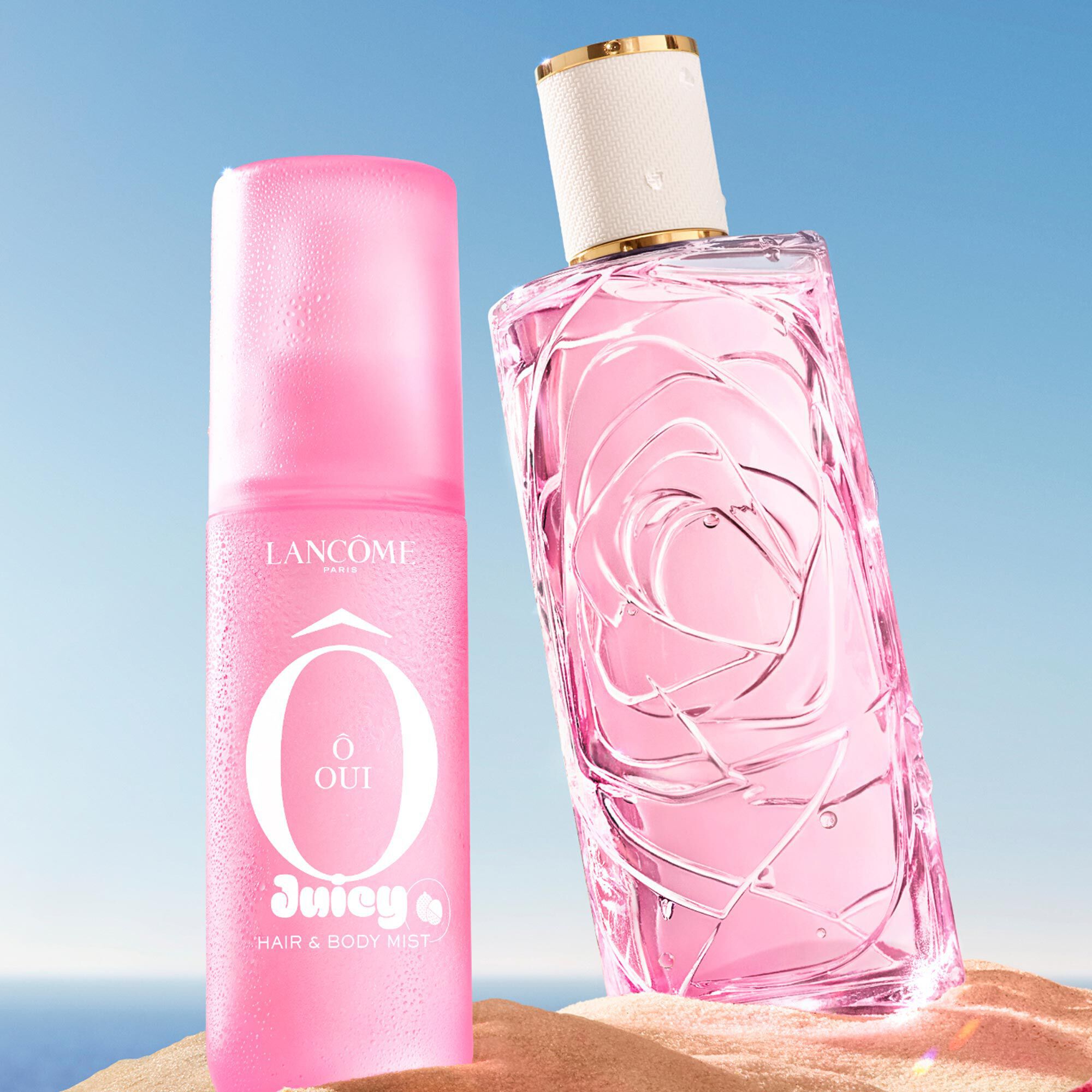 Two pink bottles, one perfume rectangular bottle with a textured design and one spray bottle, standing on a sandy beach. A folded pink striped beach towel lies next to the bottles. In the background, there is a clear blue sky and the ocean.