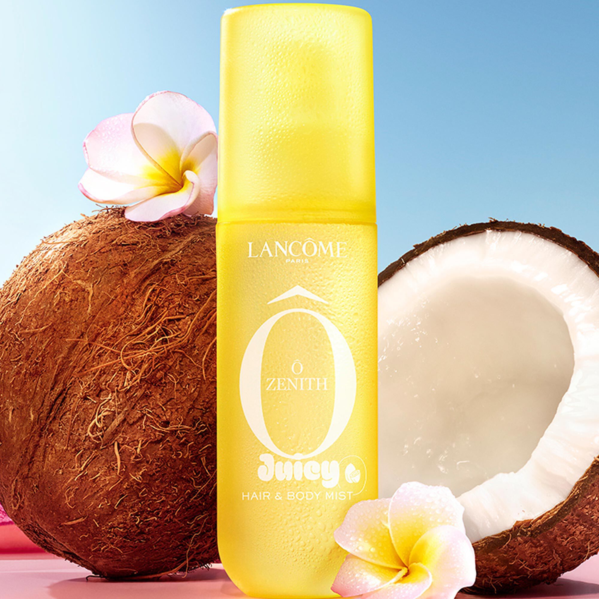 A yellow transparent spray bottle, next to coconuts and white tiare flowers, resting on a striped pink bar, with a rolled pink and white striped towel in the background, against a clear blue sky.