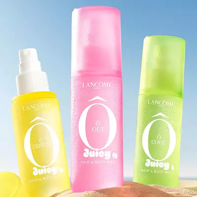 Two pink bottles, one perfume rectangular bottle with a textured design and one spray bottle, standing on a sandy beach. A folded pink striped beach towel lies next to the bottles. In the background, there is a clear blue sky and the ocean.