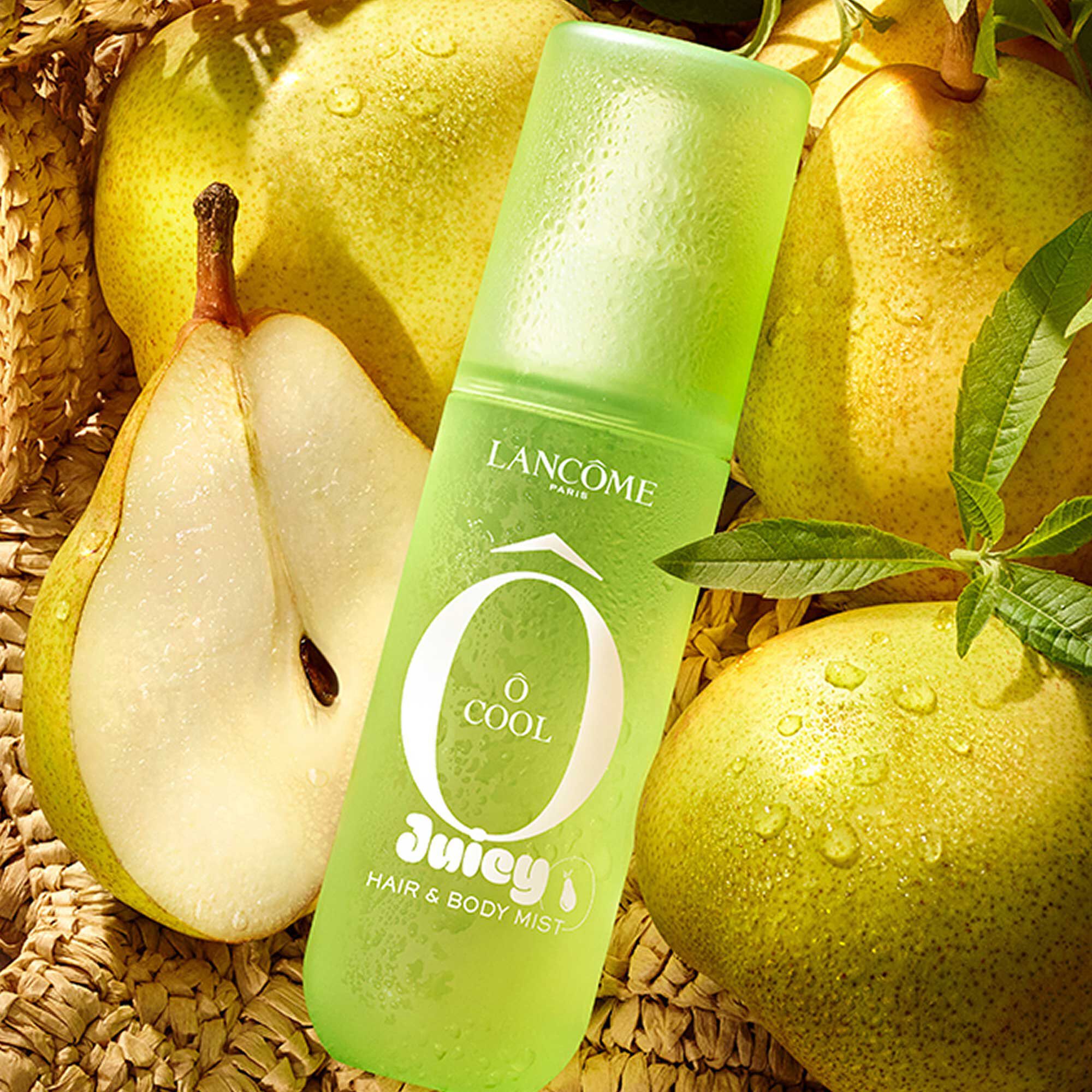 A green spray bottle in a woven bag filled with green pears and verbena branches, sits on light sand next to a pink striped towel.