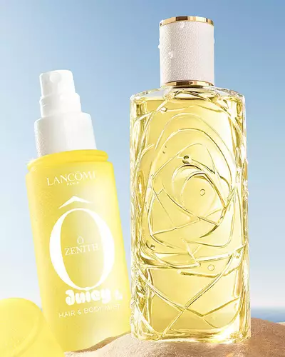 Three colorful spray bottles, one yellow, one pink, and one green, standing on a sandy beach next to a pink striped towel. The ocean is in the background under a clear blue sky.