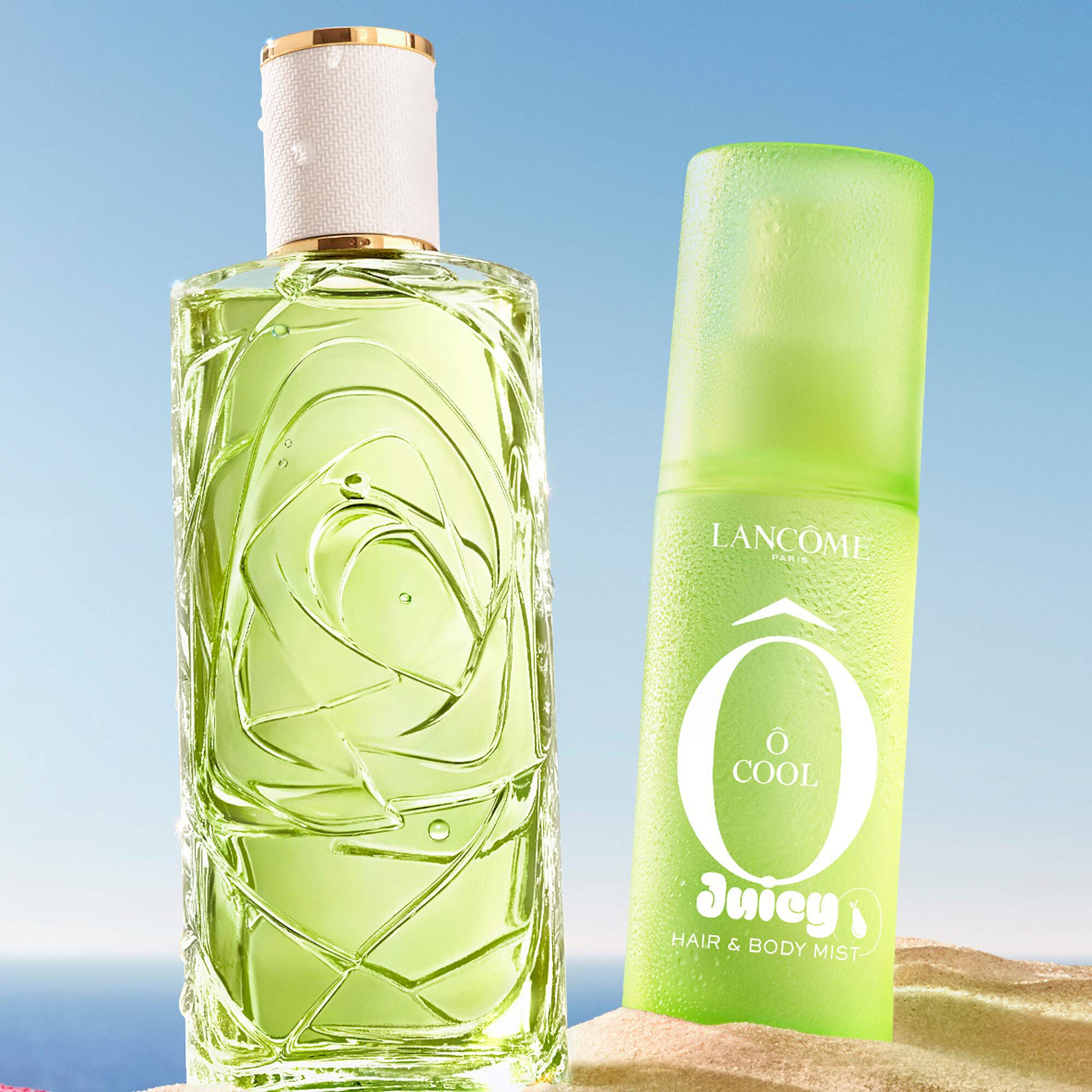 Two green bottles, one perfume rectangular bottle with a textured design and one spray bottle, standing on a sandy beach. A folded pink striped beach towel lies next to the bottles. In the background, there is a clear blue sky and the ocean.
