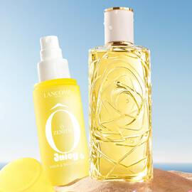 Two yellow bottles, one perfume rectangular bottle with a textured design and one spray bottle with a yellow cap on the side, standing on a sandy beach. A folded pink striped beach towel lies next to the bottles. In the background, there is a clear blue sky and the ocean.