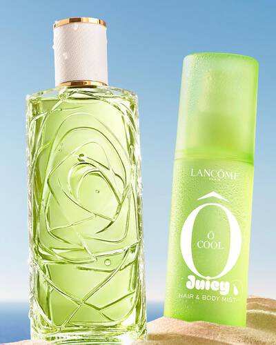 Three colorful spray bottles, one yellow, one pink, and one green, standing on a sandy beach next to a pink striped towel. The ocean is in the background under a clear blue sky.