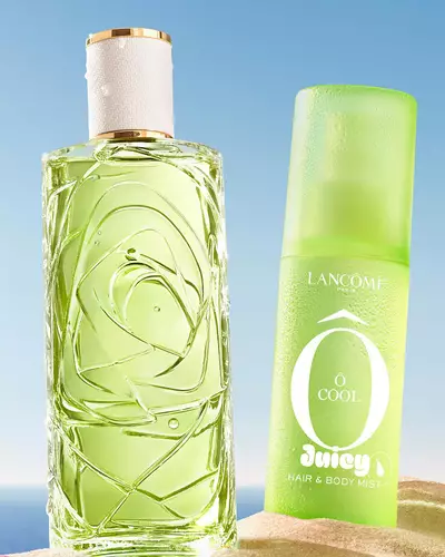 Three colorful spray bottles, one yellow, one pink, and one green, standing on a sandy beach next to a pink striped towel. The ocean is in the background under a clear blue sky.