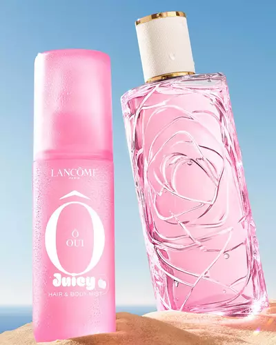 Three colorful spray bottles, one yellow, one pink, and one green, standing on a sandy beach next to a pink striped towel. The ocean is in the background under a clear blue sky.